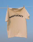 A cream-colored t-shirt hanging from a clothesline with wooden clothespins against a clear blue sky and sea background. The word 'NANTUCKET' is printed in black arched letters across the chest.