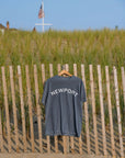 A faded blue-gray t-shirt with 'NEWPORT' printed in white arched text across the chest, hanging on a wooden hanger from a wooden beach sand fence with sand dunes and an American flag in the background.