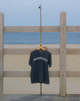 A dark navy blue t-shirt with 'NEWPORT' printed in white across the chest, hanging on a wooden hanger from a beachside metal shower stand. The background features a sandy beach, blue ocean, and a light-colored wooden pier railing.