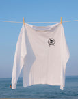 A white long-sleeve t-shirt with a black circular graphic logo on the left chest, hanging from a clothesline with wooden clips against a blue sky and ocean background.