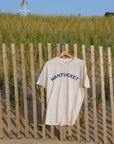 An off-white short-sleeve t-shirt with 'NANTUCKET' printed in navy blue arched text across the chest, hanging on a wooden hanger from a wooden slat fence at a beach with sand dunes and grass in the background.