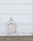 A cream-colored sweatshirt with 'NANTUCKET' printed in blue on the chest, hanging on a wooden hanger against a white garage door.